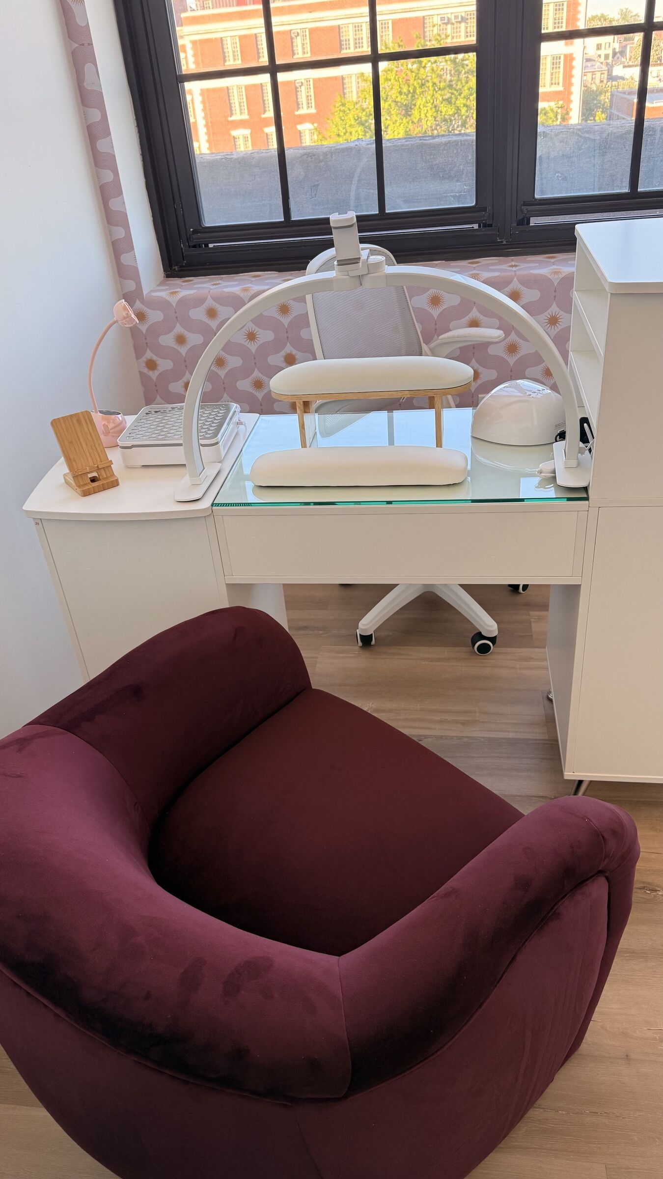 Nail Studio detail: manicure desk with white LED arch lamp, gel dryer, dust extractor, and pink-orange accent wallpaper with daylight from industrial window.