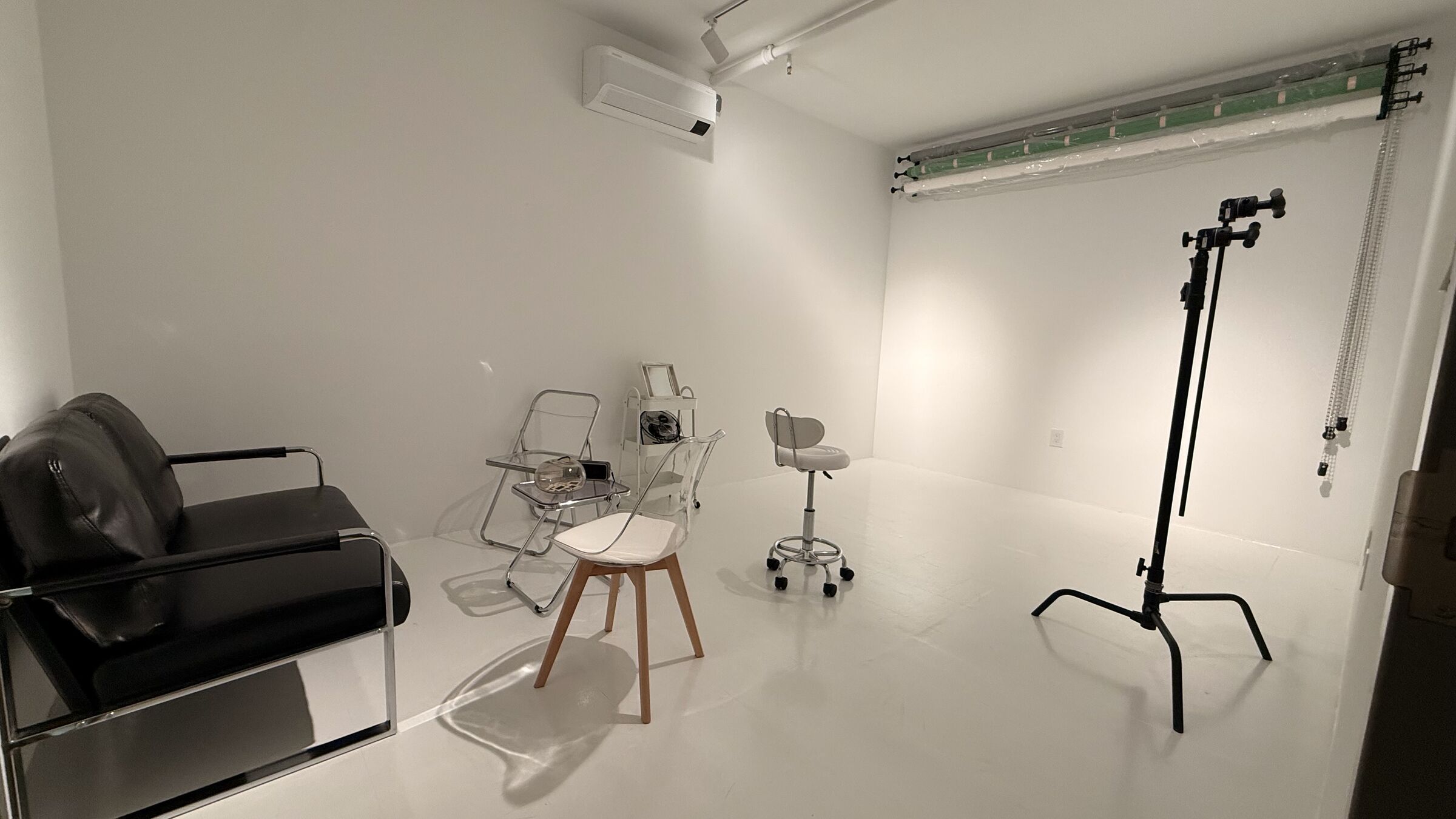 Portrait Studio in Sunset Park — white seamless backdrop rolls on the back wall, C-stand and Nanlite LED key, black leather couch and clear acrylic chairs, white-painted wood floor.