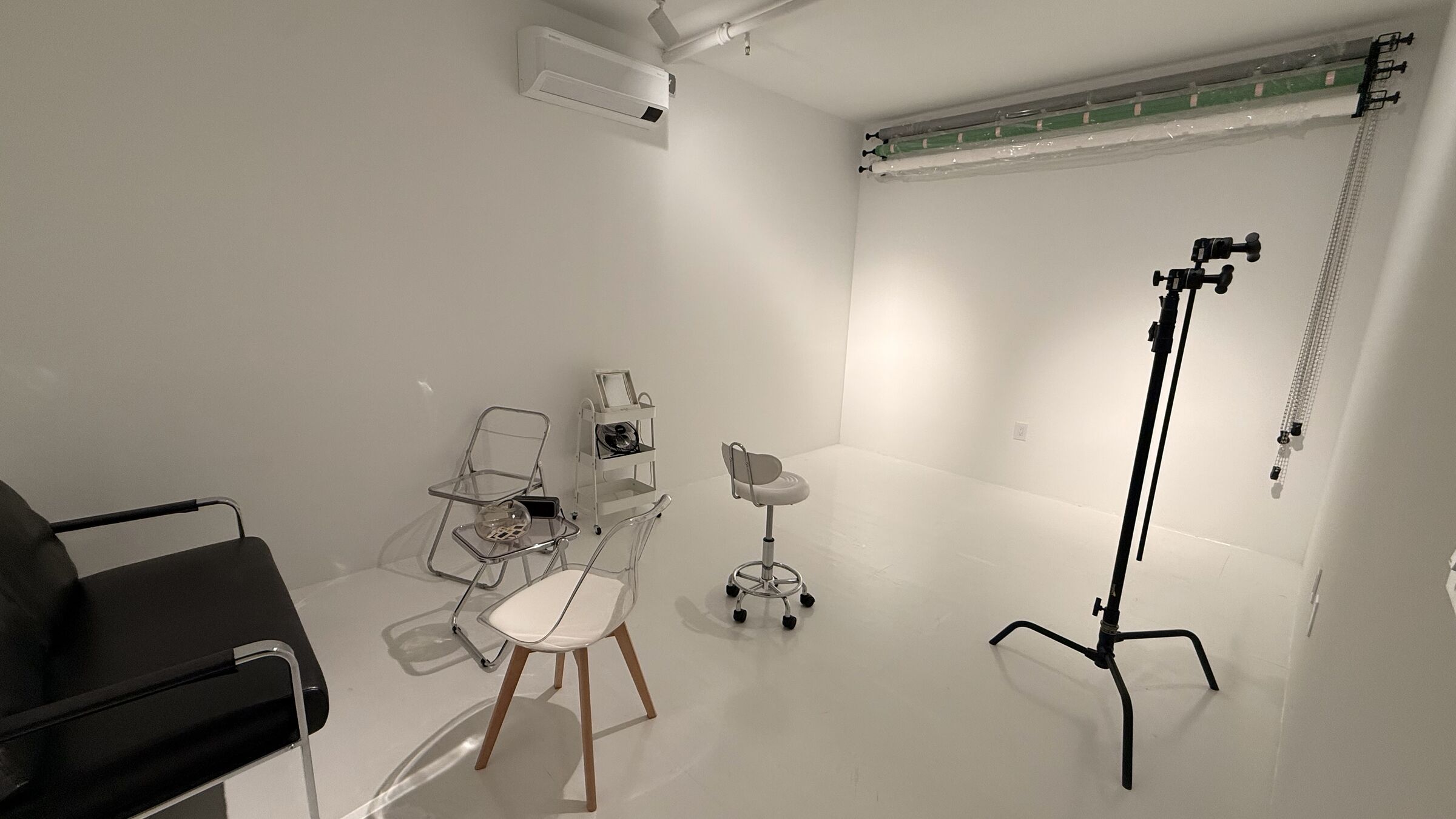 Portrait Studio: full room scale with C-stand prominent, seamless backdrop rolls on the back wall, black leather couch and clear acrylic chairs foregrounded.